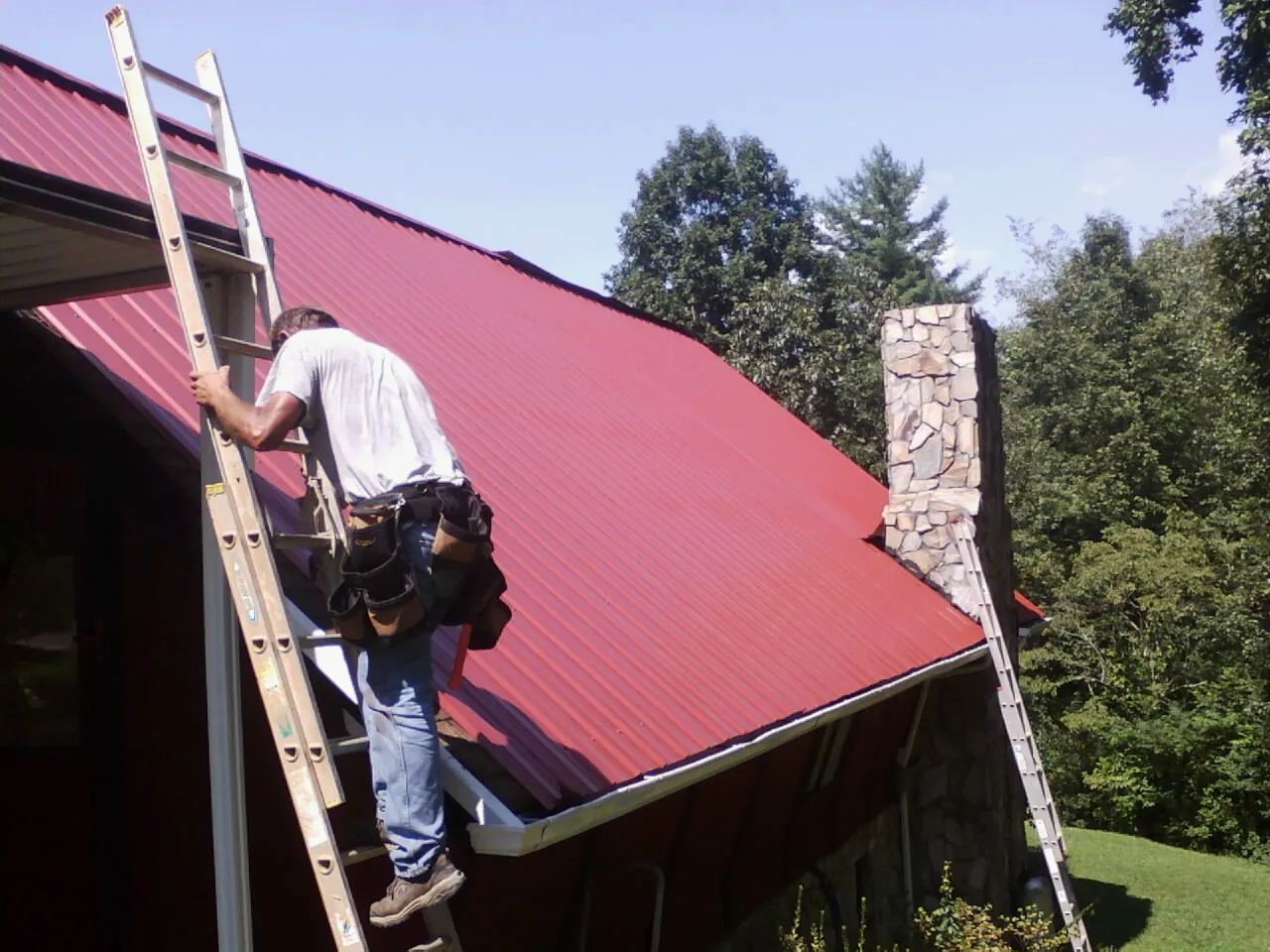 Roofer on a ladder beside a red metal roof and stone chimney — Soffit & Fascia Repair by Roofing Brothers in Cooleemee